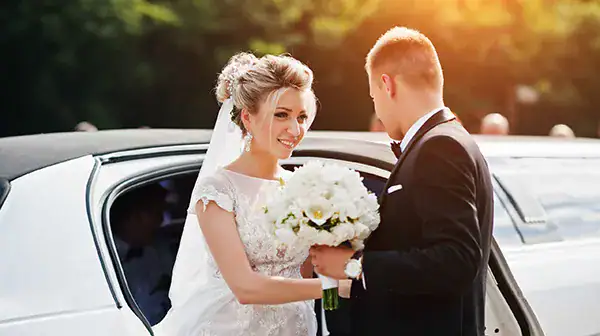 A person in a white dress holding a bouquet stands beside a white car, greeted by another individual in a black suit, with sunlight filtering through surrounding greenery. This joyful wedding moment reflects the elegance and celebration often seen in Atlantic City, NJ.