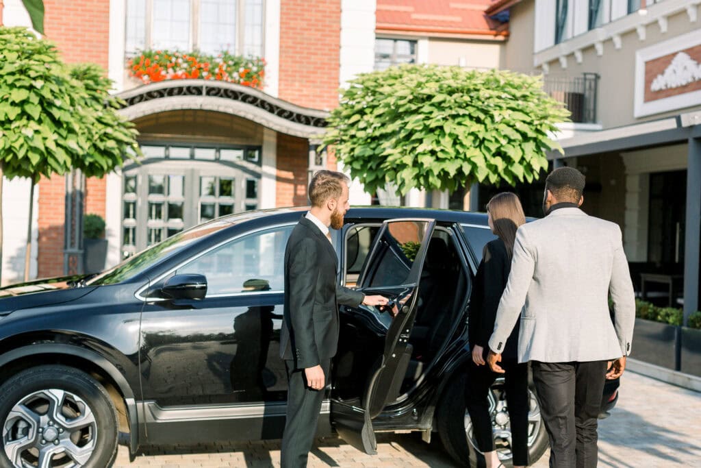 A valet in a black suit opens the door of a black SUV from a limo service Atlantic City for a man and woman in business attire outside a stylish NJ building with trees and large windows.