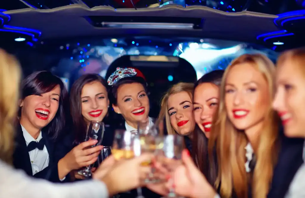 A group of smiling women, dressed up and sitting in a limousine, holding and clinking glasses in celebration, are ready for a fun night out in Atlantic City, NJ. The atmosphere is festive with blue lights and a joyful mood as they head to enjoy the casinos and boardwalk of the Jersey Shore.