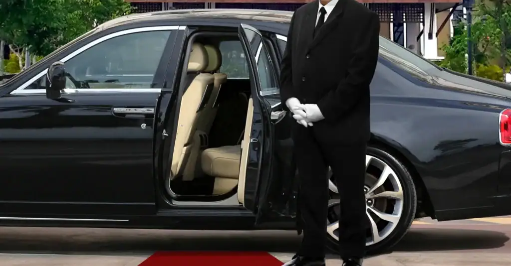 A chauffeur in a black suit and white gloves stands next to a luxury black car with the rear door open, ready for a passenger, on a red carpet. This elegant scene could be taking place right outside one of the high-end resorts in Atlantic City, NJ